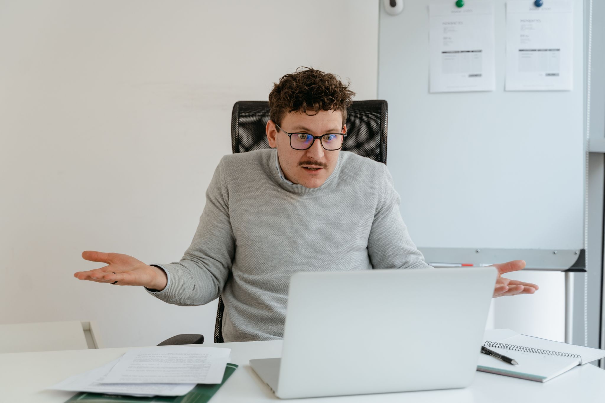 A man in a gray sweater and glasses gesturing at his laptop screen during a video call in an office setting.
