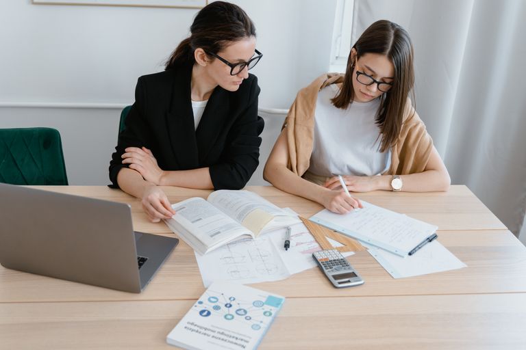 Two women at a desk studying with books, notes, and a laptop, emphasizing collaboration.
